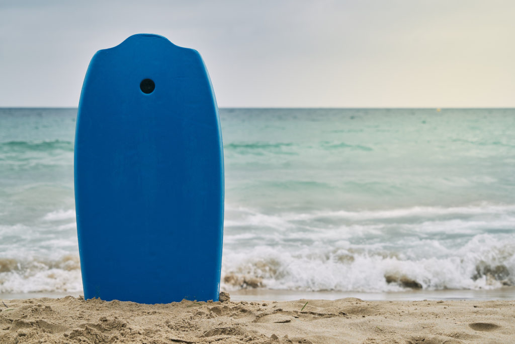 A closeup of a blue boogie board in an upright position on a sandy shore with beach waves in the background
