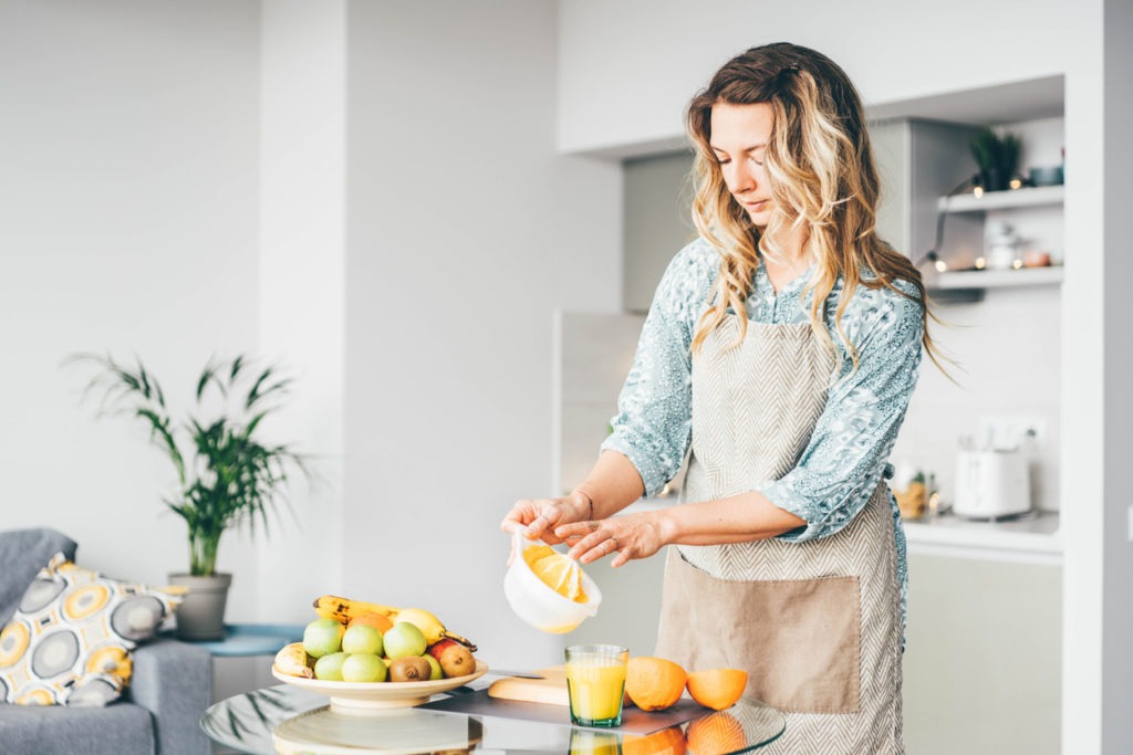 Pretty curly-haired lady in grey apron pours fresh orange juice into the cup from squeezer at the glass table in the light contemporary kitchen in the morning