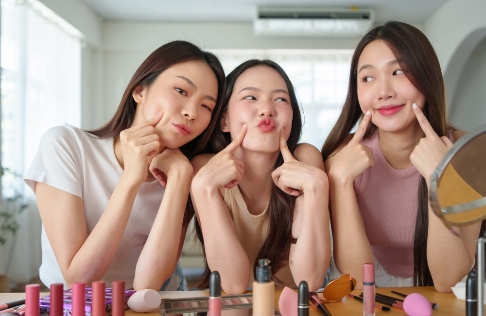 Three young Asian women having fun with makeup, Happy Korean girls making playful faces in a beauty setup with cosmetics on table, expressing joy and friendship
