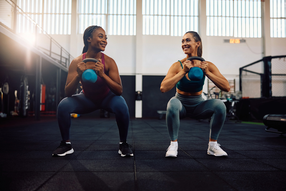 Young happy sportswomen using kettle bells while exercising squats in health club. Copy space