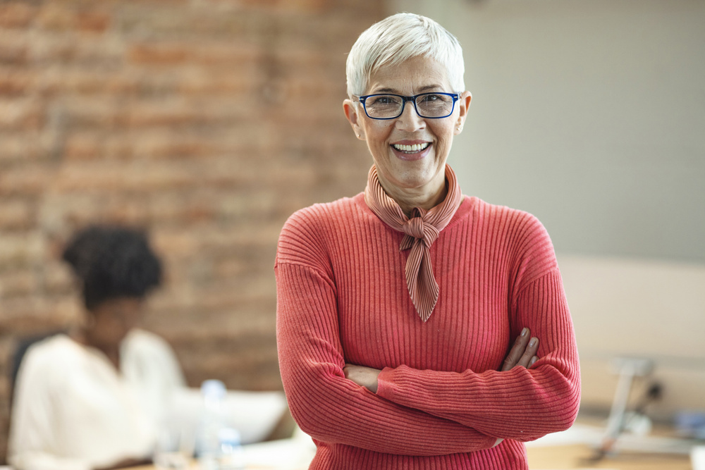 Close-up portrait of mature business woman standing at office. Pretty older business woman, successful confidence with arms crossed in financial building. Mature female in office with team meeting in backgroundClose-up portrait of mature business woman standing at office. Pretty older business woman, successful confidence with arms crossed in financial building. Mature female in office with team meeting in background