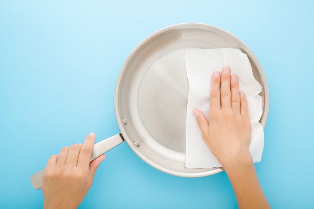 On a table with a light blue background, a young adult woman is scrubbing a stainless steel frying pan with a dry white paper napkin. 