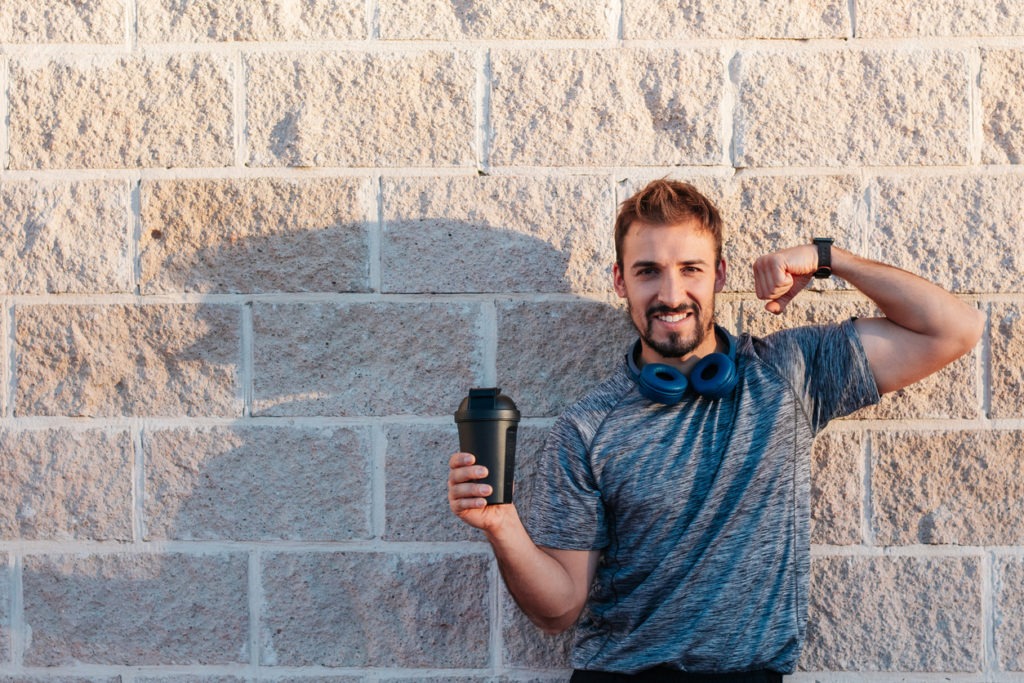 A man holding a bottle blender, smiling and showing off his arm muscles