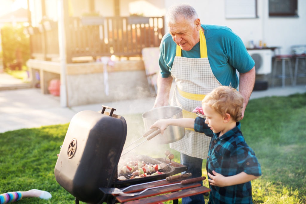 Proud grandfather is carefully teaching his young adorable boy how to grill