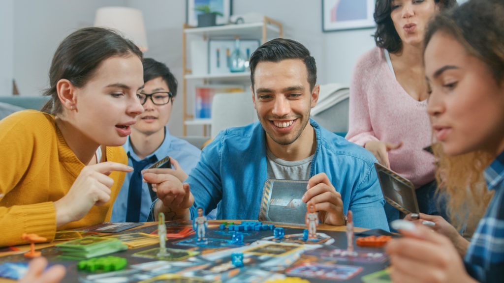 Diverse Group of Guys and Girls Playing in a Strategic Uniquely Designed Board Game with Cards and Dice. Friends Having Fun Reading Cards, Joking, Making Moves, and Laughing in a Cozy Living Room