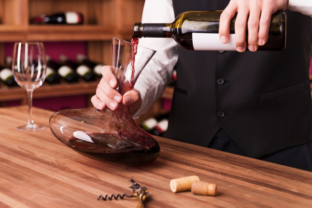 Confident male sommelier pouring wine to decanter while standing near the wine shelf