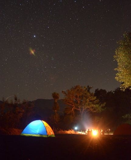 Glowing yellow tent and a bonfire surrounded by campers under a starry night sky