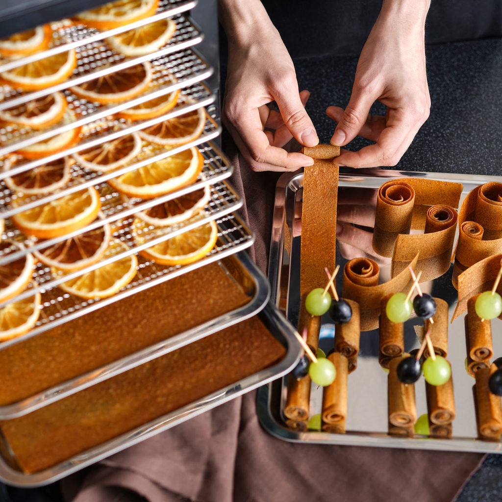 Male hands twisting orange pastille and coocing canapes. Dehydrator with trays for drying fruits and vegetables. Frips and Pastille on grates