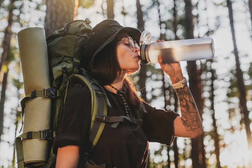 Side attractive happy young woman hiker with backpack drinking bottled water traveling in country forest
