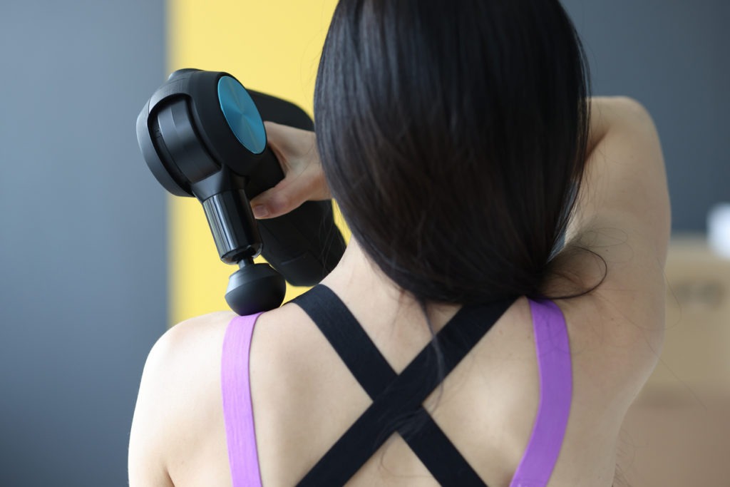 Woman doing massage of muscles of neck and back with percussion massager closeup