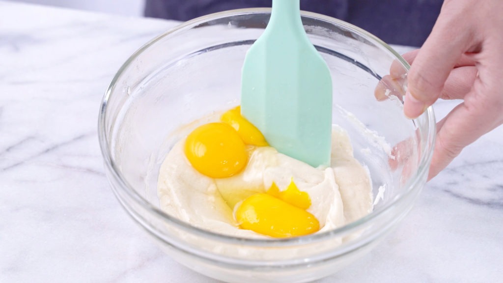 Mixing egg yolk into the cake batter with a green rubber spatula mixer tool stirring until smooth and blending well in a glass bowl, close up, lifestyle