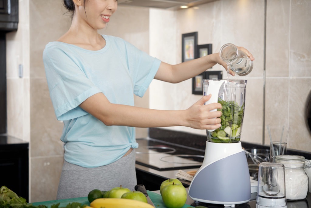 Positive fit Asian woman adding water into kitchen blender before mixing ingredients for a smoothie