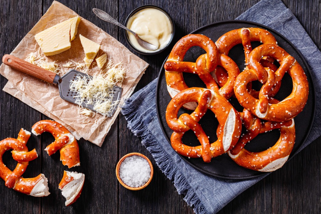 Soft pretzels baked in the form of knot and sprinkled with salt on black plate on dark wooden table with cheese sauce, grated cheese and grater.