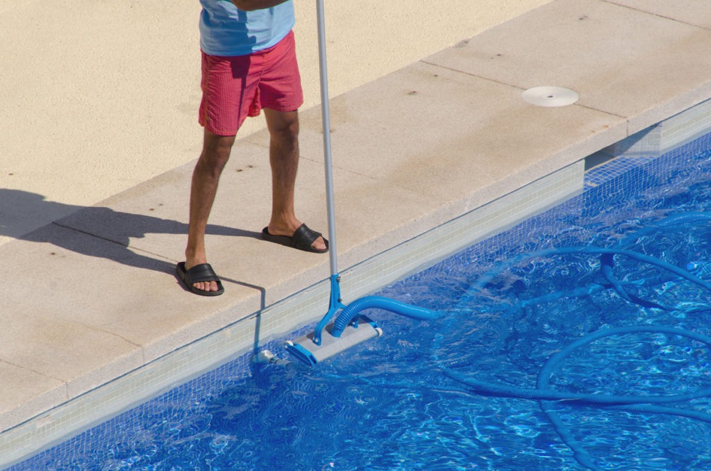 A Man cleans the swimming pool ground with a vacuum head and a telescopic pole. Summer pool maintenance service