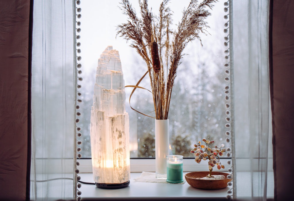 Rough big selenite crystal tower pole lamp illuminated on home window sill, spiritual home decor accent. Winter forest on background