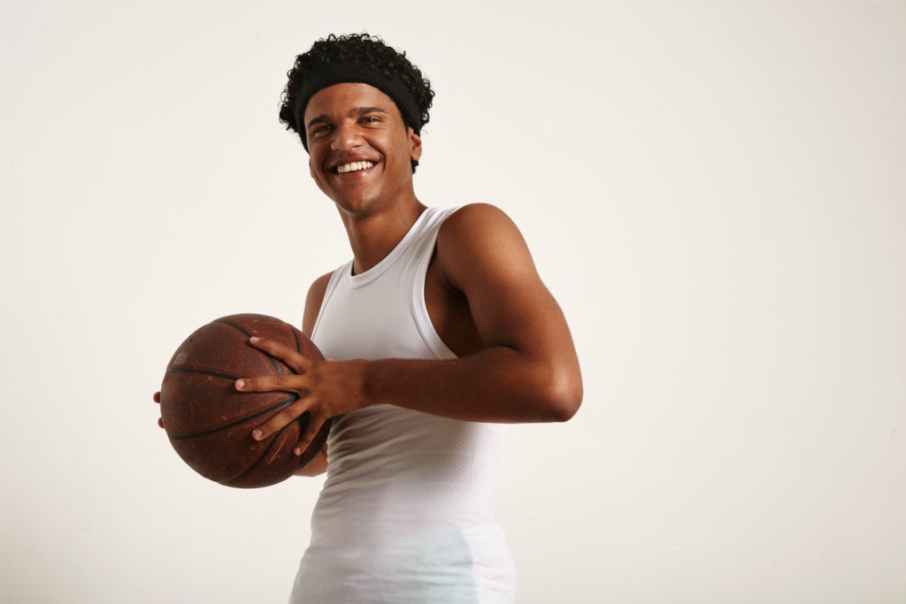 A low-angle quarter shot of a cheerful laughing young attractive African American wearing a white sleeveless shirt and a headband holding a grunge leather basketball to his chest isolated on white