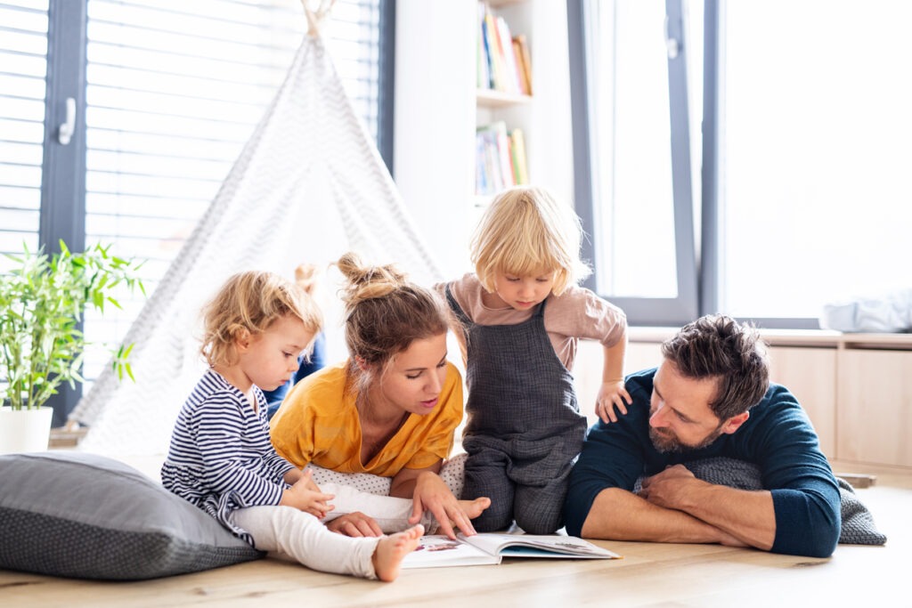 A young family with two small children in a bedroom reading a book under a tepee