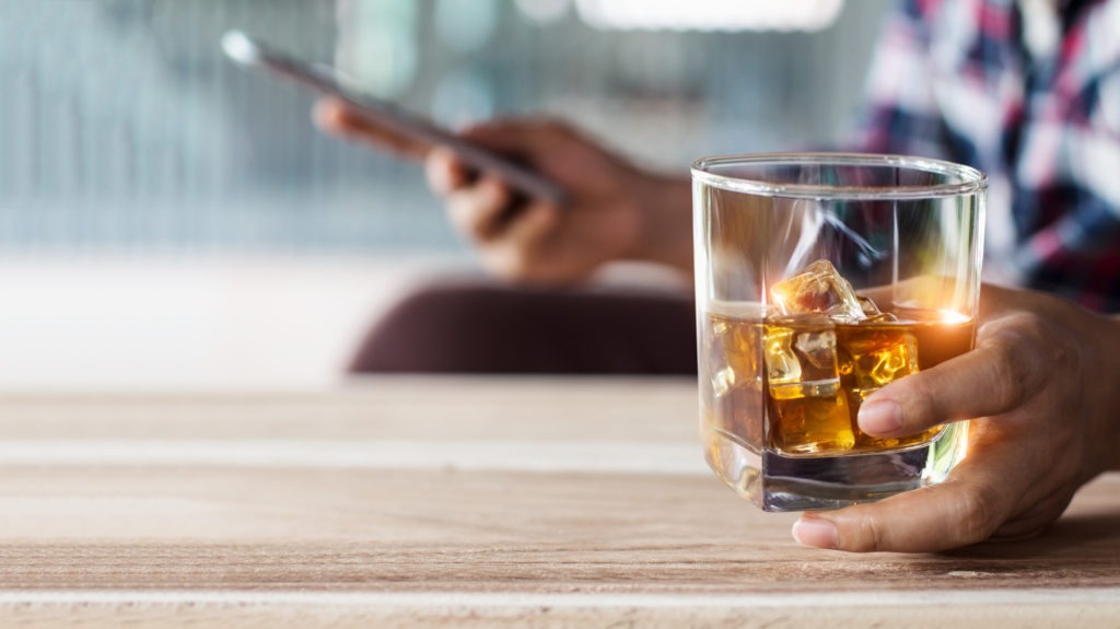 Man-relaxing-with-bourbon-whiskey-drink-alcoholic-beverage-in-hand-and-using-mobile-smartphone-chat-with-social-network-at-bar-counter-in-home-after-work
