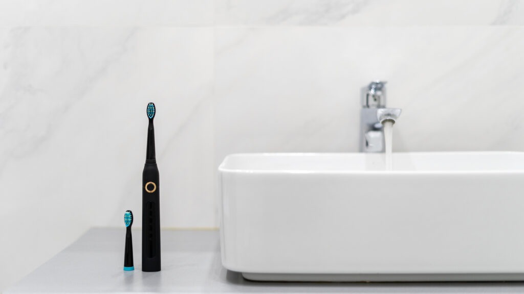Electric toothbrush with replaceable brush near white sink in bright clean bathroom with flowing faucet
