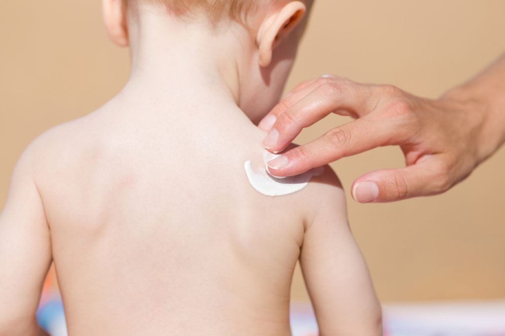 Hand of a young mother applying sunscreen lotion on baby’s shoulder
