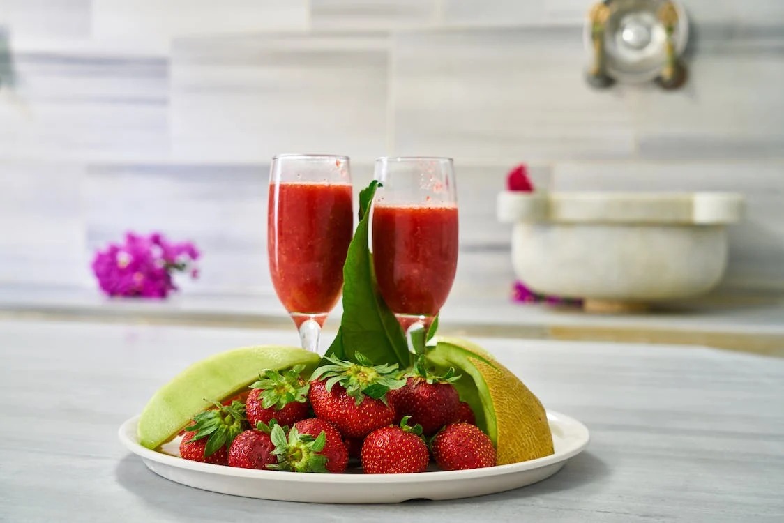 A plate of strawberries beside 2 glass of fruit juice