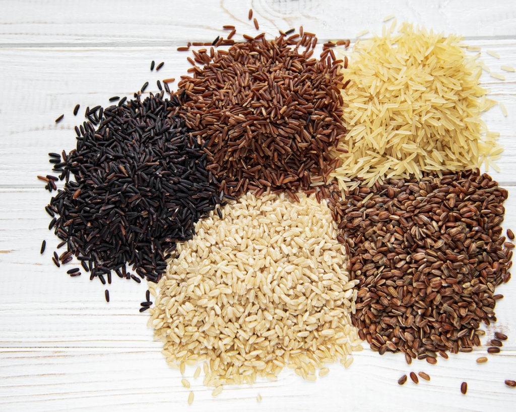Black, basmati, brown, and mixed red rice are shown in a set against a white wooden background.