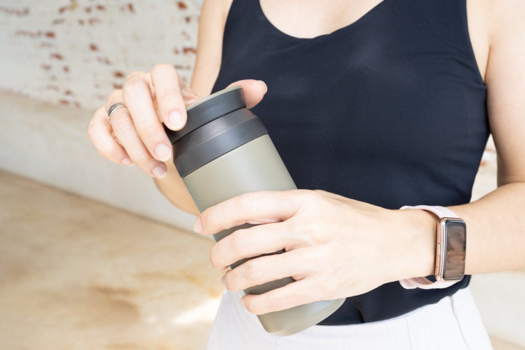 Close up of woman's hand wearing a black tank top, holding a green reusable insulated water bottle and about to open the bottle to drink after exercise