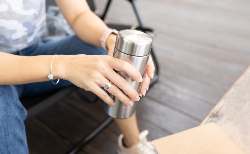 Woman is sitting at an outdoor cafe, holding stainless steel reusable insulated water bottle relaxing)