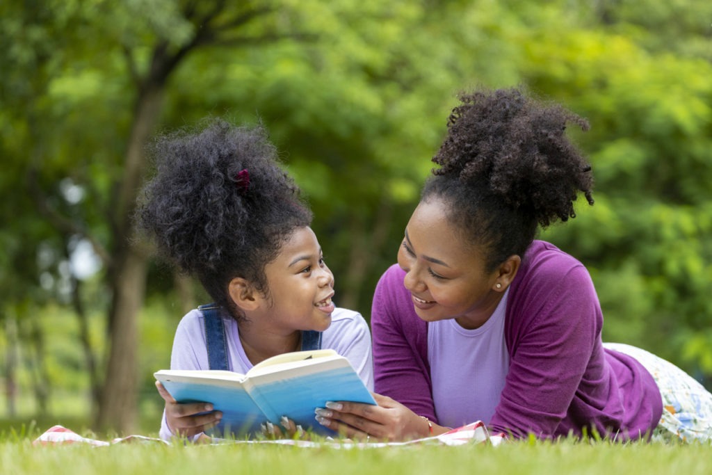 African mother is teaching her young daughter to read while lying down after having a summer picnic in the public park for education and happiness concept