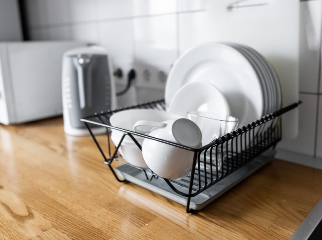Dish rack holds many dishes and cups against wooden countertop, white wall tiles, sink and faucet. Budget and lightweight antimicrobial dish drainer with drain board at modern scandinavian kitchen