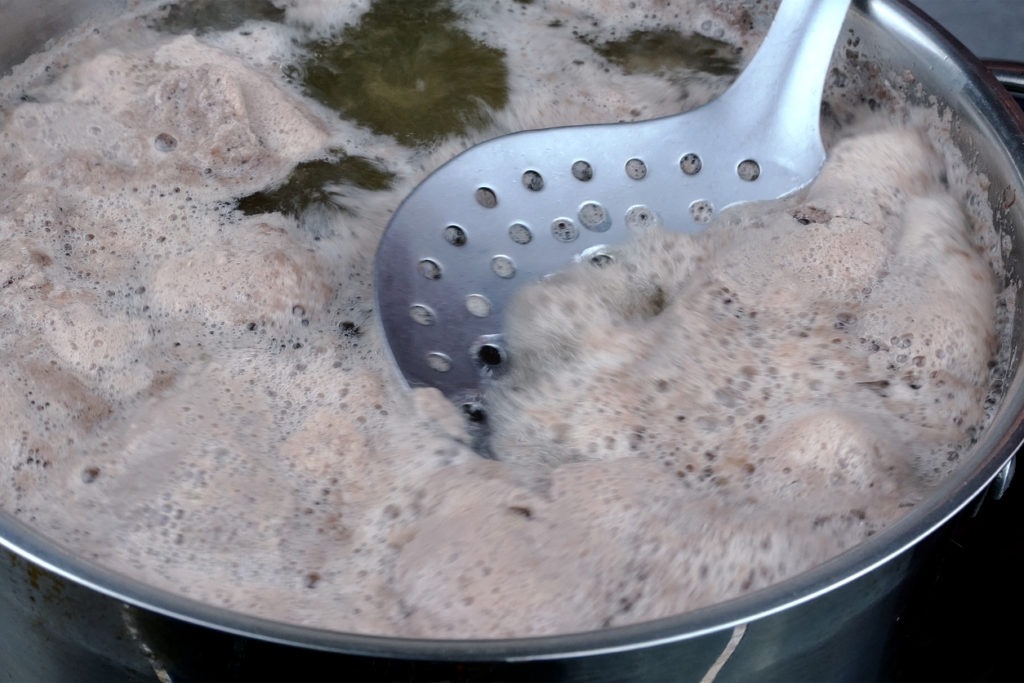 Meat broth with abundant foam boils in saucepan, foam is removed with metal slotted spoon.