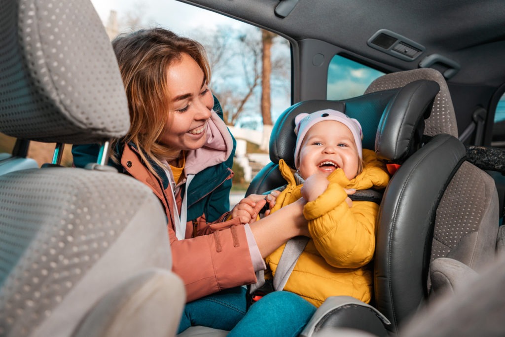 Smiling caucasian mother prepares her little cute child for a trip by car. A woman fastens her seat belts in a child seat. View inside a car. Family autumn vacation