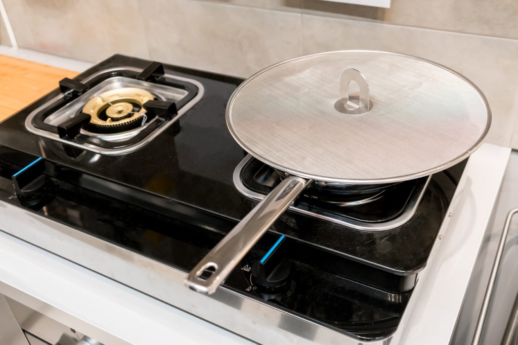Silver metal frying pan covered with oil splatter screen on gas stove