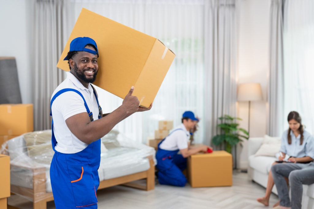 Smiling man mover worker in blue uniform carrying cardboard boxes moving to living room in new house.Professional delivery and moving service.