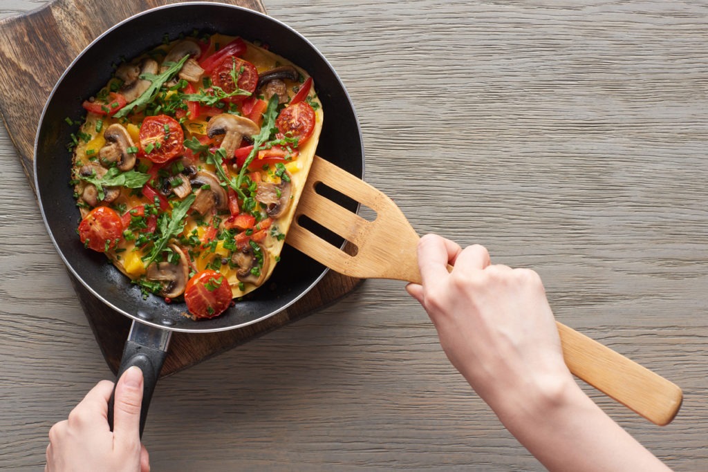 woman cooking omelet with mushrooms, tomatoes and greens on frying pan