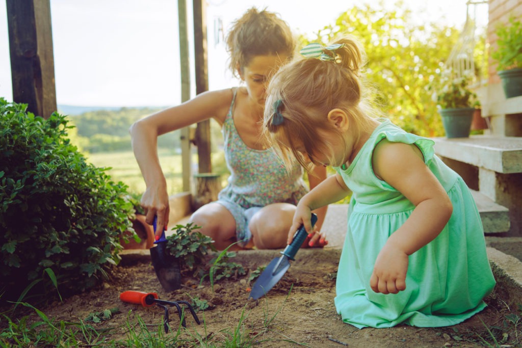 Young mother and daughter digging picking ground dirt land in their yard garden near the home house family planting flowers or seeds using gardening tools in summer day