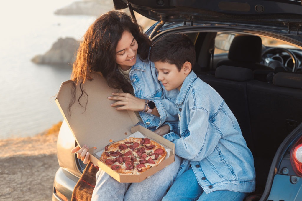 A mother holding a box of pizza while the son looks on.