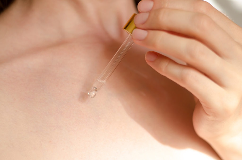 A pipette in a woman's hand dribbles serum or essential oil onto her body. 