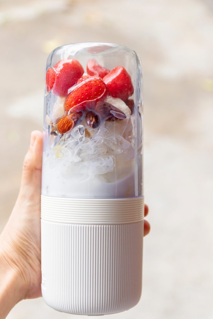 A woman holding a portable blender with strawberries, banana, almond, milk, and ice