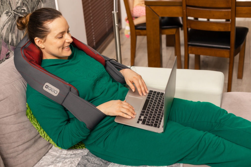 A woman using a neck and shoulder massager while working on a laptop