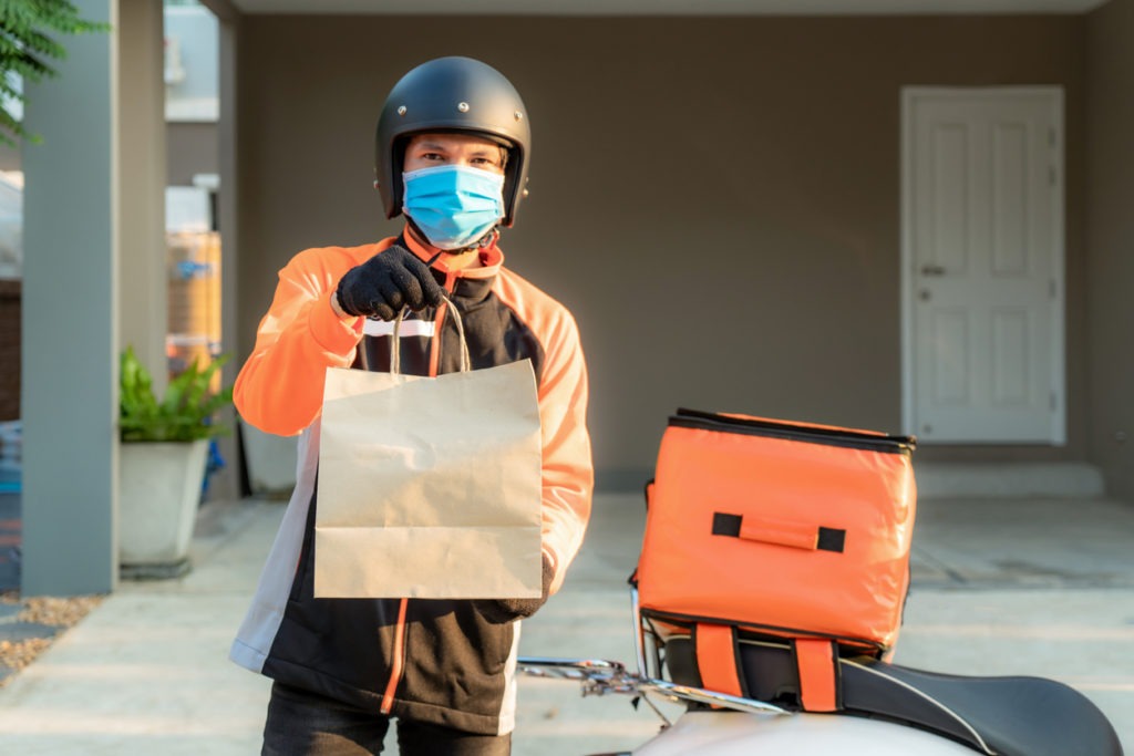 Delivery Asian man wearing a protective mask in an orange uniform and is ready to send delivery. Food bag in front of customer house with case box on a scooter, express food delivery, and shopping online concept