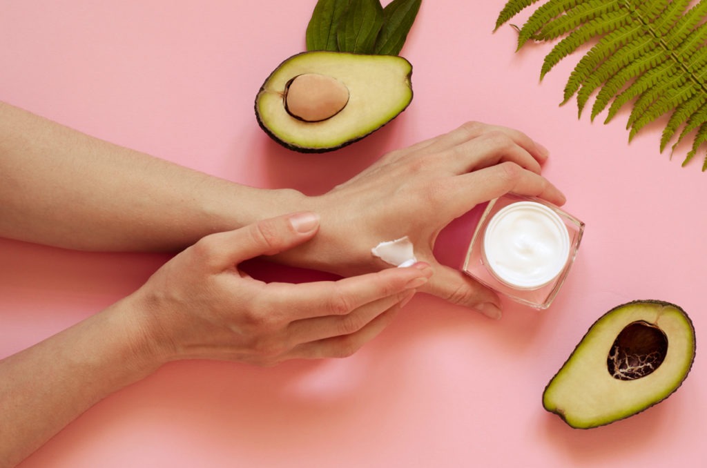 Girl spreads cream on her hands. On a pink background, there are two avocado halves with leaves, a bowl with avocado oil, a jug of cream, and a fern leaf.