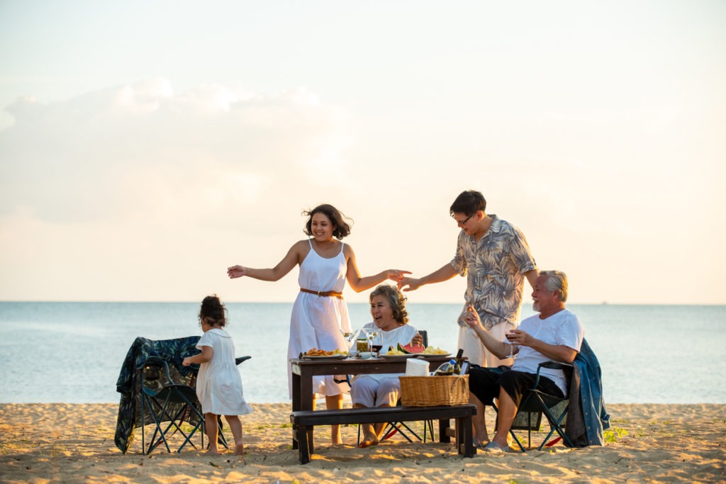 Happy Asian family on holiday vacation. Group of a multi-generation family little girl with parents and grandparents relax and enjoy a dinner party and dancing together on the beach at summer sunset