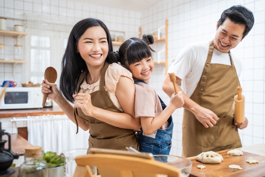 Happy Asian family stays at home in the white-colored kitchen to bake and dance together.