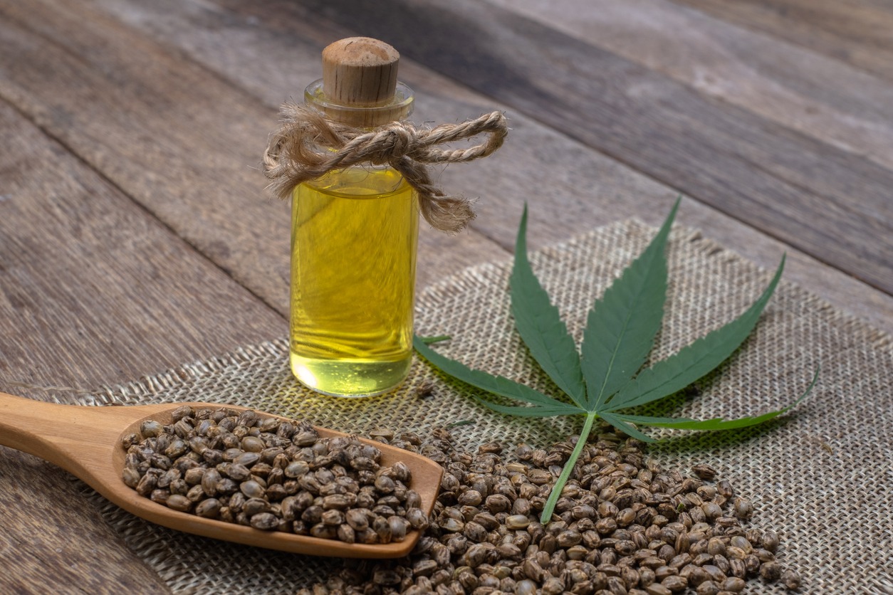 Hemp seeds and a leaf are all around a glass container of hemp oil that is bow-tied