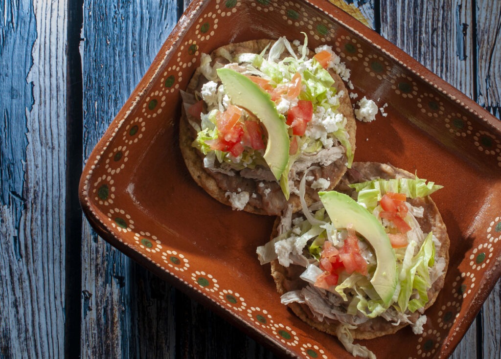 Tostadas, a traditional Mexican dish, are served with chicken or carnitas, lettuce, tomato, onion, fried beans, hot red salsa, and cream over a rustic table.