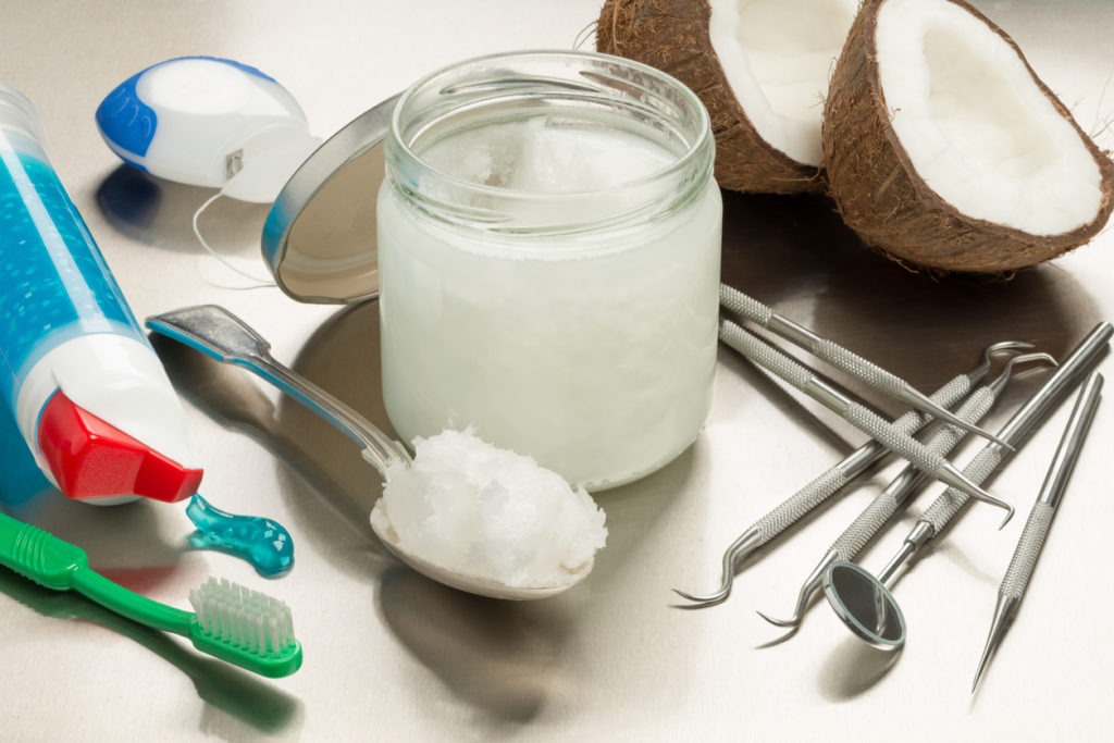 Selection of dental instruments with coconut oil, toothpaste, a toothbrush, and floss against a stainless steel background.