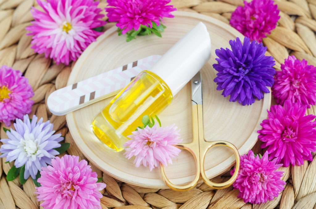 Small bottle of cosmetic oil, scissors and nail file placed on a white ceramic surrounded by flowers. 