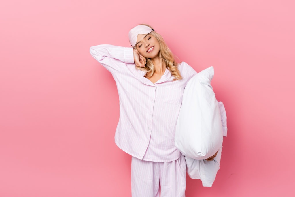 Smiling woman in pajamas and blindfold holding a pillow and stretching on pink background