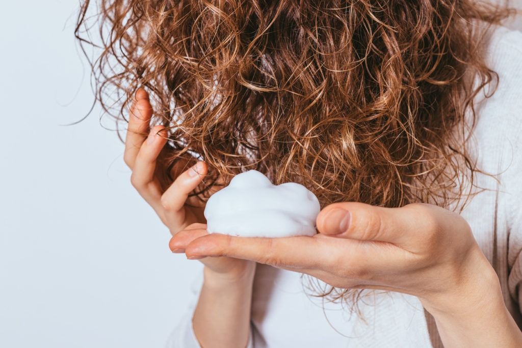 Woman applying styling mousse to her curly hair in close-up against a white studio background.
